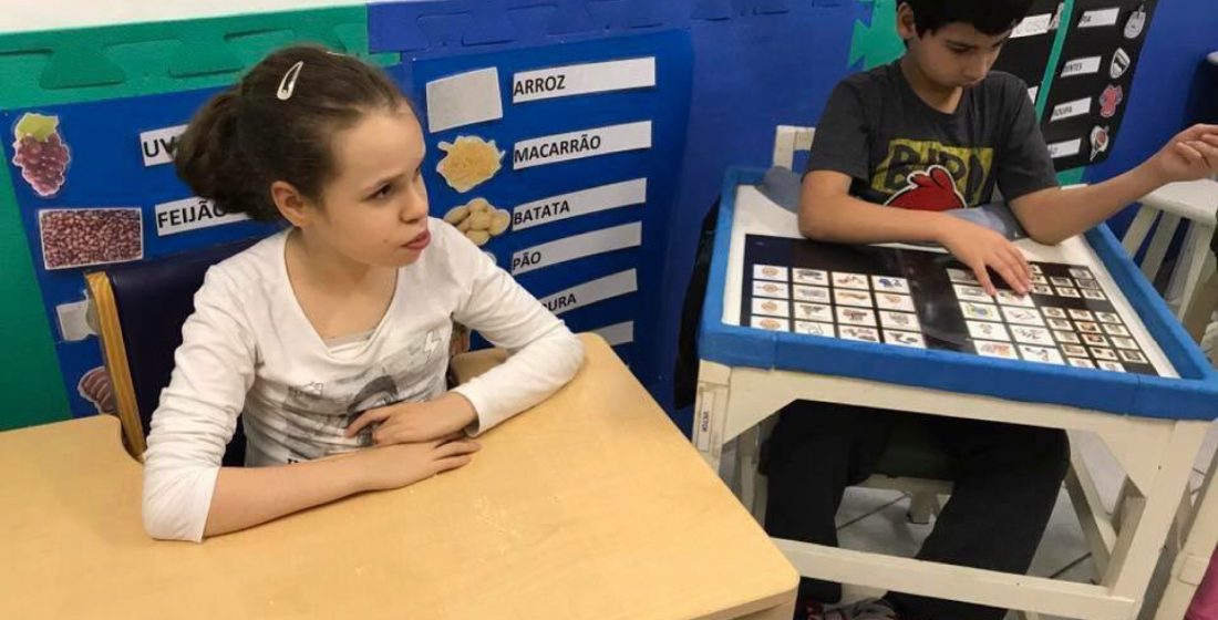 duas crianças sentadas em uma mesa escolar adaptada dentro de uma sala de aula